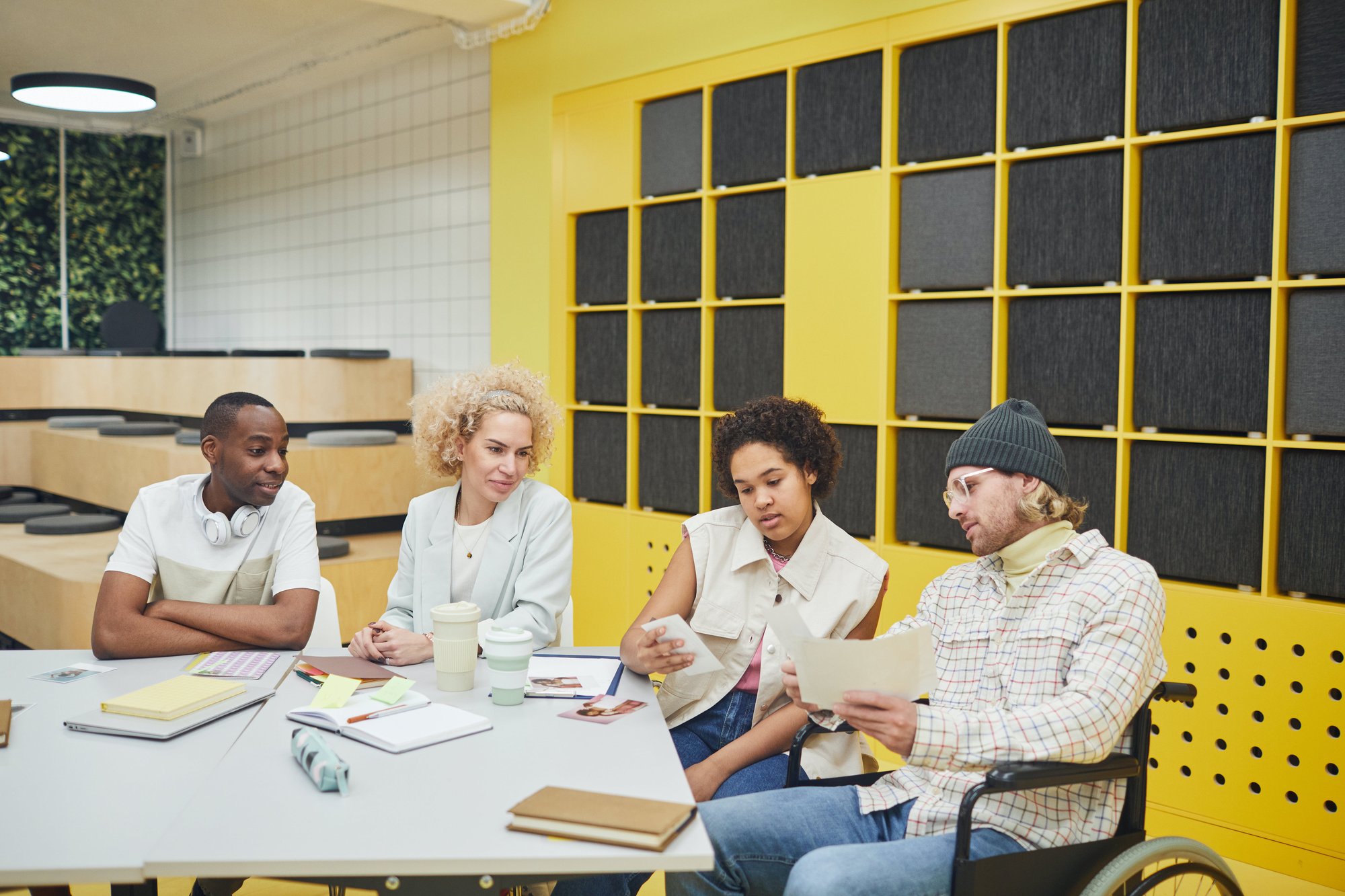 Four people are sitting at a table having a discussion with notebooks, coffee cups, and documents in front of them. Four people are sitting at a table having a discussion with notebooks, coffee cups, and documents in front of them.